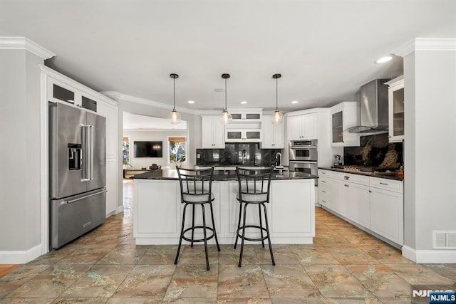 kitchen featuring wall chimney range hood, appliances with stainless steel finishes, white cabinetry, a kitchen breakfast bar, and a center island with sink