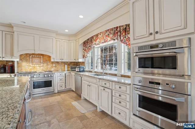 kitchen featuring appliances with stainless steel finishes, sink, decorative backsplash, light stone countertops, and cream cabinetry