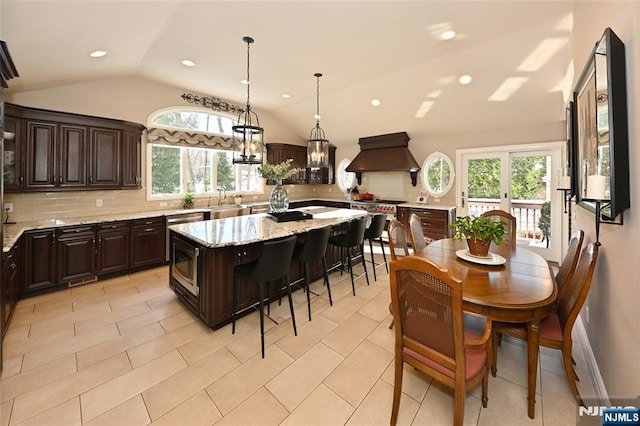 kitchen featuring a kitchen island, lofted ceiling, dark brown cabinets, light stone counters, and custom range hood