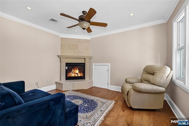 living room with a tiled fireplace, wood-type flooring, ornamental molding, and ceiling fan