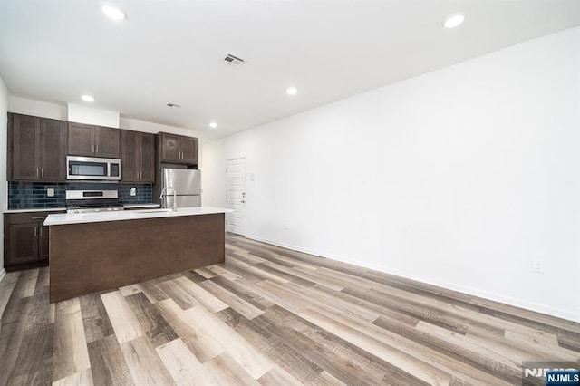 kitchen featuring a kitchen island with sink, dark brown cabinetry, appliances with stainless steel finishes, light countertops, and decorative backsplash