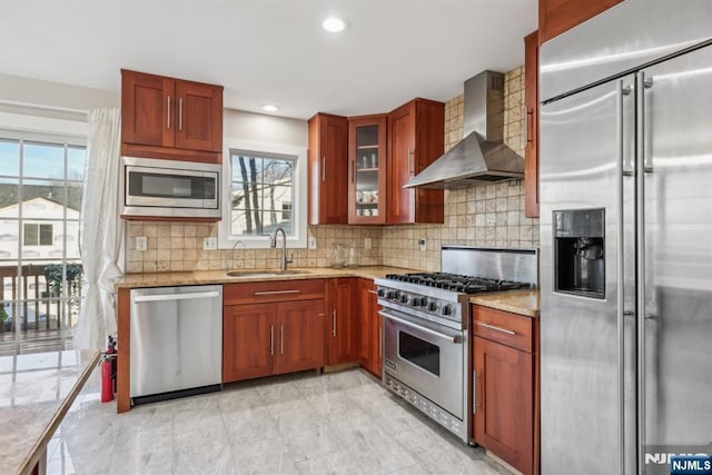 kitchen featuring wall chimney range hood, sink, built in appliances, light stone counters, and tasteful backsplash