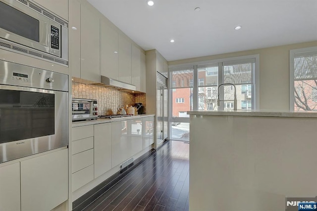 kitchen featuring appliances with stainless steel finishes, white cabinetry, decorative backsplash, exhaust hood, and dark wood-type flooring