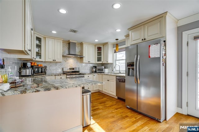 kitchen featuring decorative light fixtures, appliances with stainless steel finishes, wall chimney range hood, light hardwood / wood-style floors, and cream cabinetry