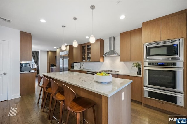 kitchen with wall chimney range hood, dark wood-type flooring, a center island, built in appliances, and decorative light fixtures