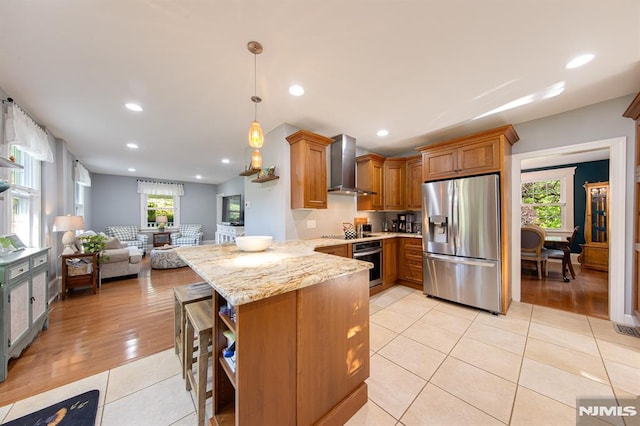 kitchen with appliances with stainless steel finishes, kitchen peninsula, pendant lighting, light stone countertops, and wall chimney range hood