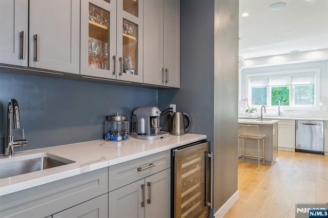 bar featuring dishwasher, sink, beverage cooler, light stone counters, and light hardwood / wood-style flooring