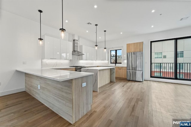 kitchen featuring appliances with stainless steel finishes, white cabinetry, hanging light fixtures, kitchen peninsula, and wall chimney exhaust hood