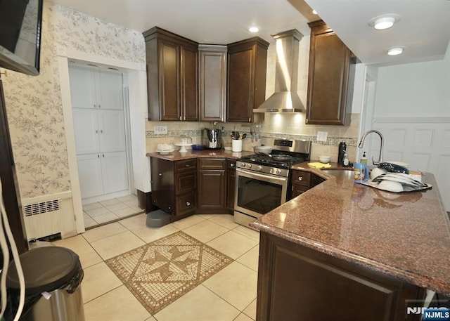 kitchen featuring radiator, light tile patterned floors, stainless steel gas range, dark stone counters, and wall chimney exhaust hood