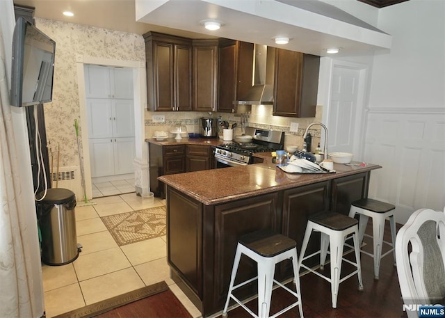 kitchen with light tile patterned floors, kitchen peninsula, gas stove, dark brown cabinets, and wall chimney range hood