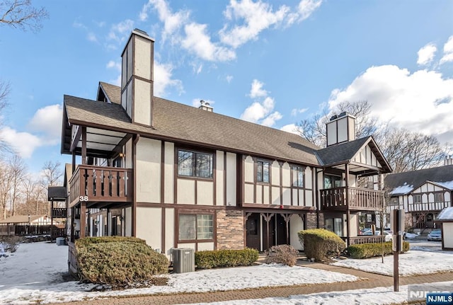 snow covered back of property featuring roof with shingles, a chimney, stucco siding, central AC unit, and a balcony