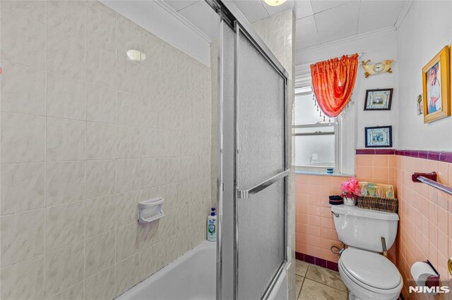 bathroom featuring tile patterned flooring, combined bath / shower with glass door, tile walls, and toilet