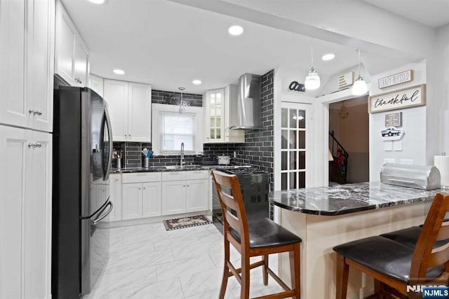 kitchen featuring stainless steel refrigerator, decorative light fixtures, a breakfast bar area, white cabinets, and wall chimney range hood