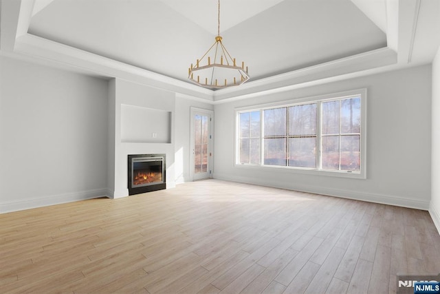 unfurnished living room featuring light wood finished floors, a warm lit fireplace, baseboards, a raised ceiling, and an inviting chandelier