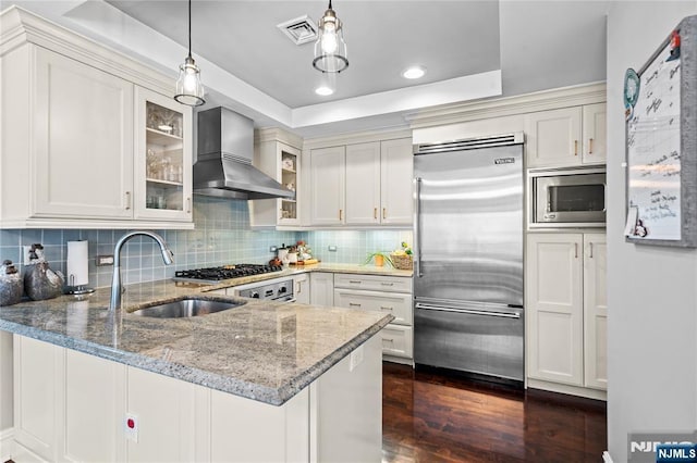 kitchen featuring kitchen peninsula, sink, built in appliances, a tray ceiling, and wall chimney exhaust hood