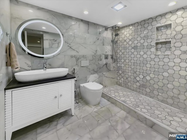 bathroom with a tile shower, vanity, tile walls, and toilet