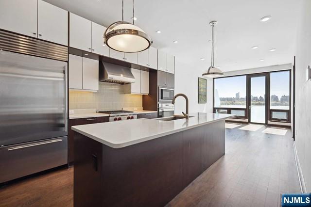 kitchen featuring white cabinetry, built in appliances, pendant lighting, a kitchen island with sink, and exhaust hood