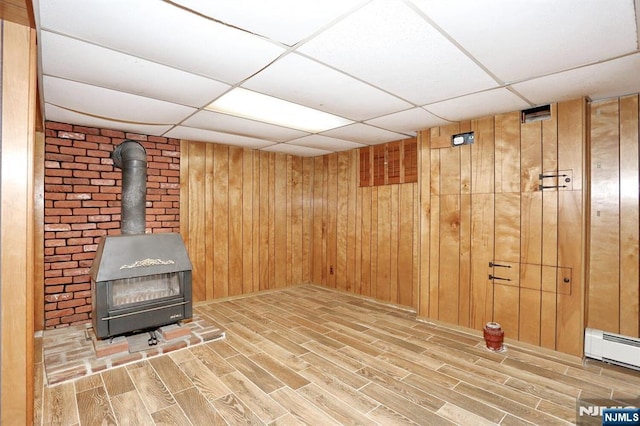 basement featuring baseboard heating, a paneled ceiling, wooden walls, and a wood stove