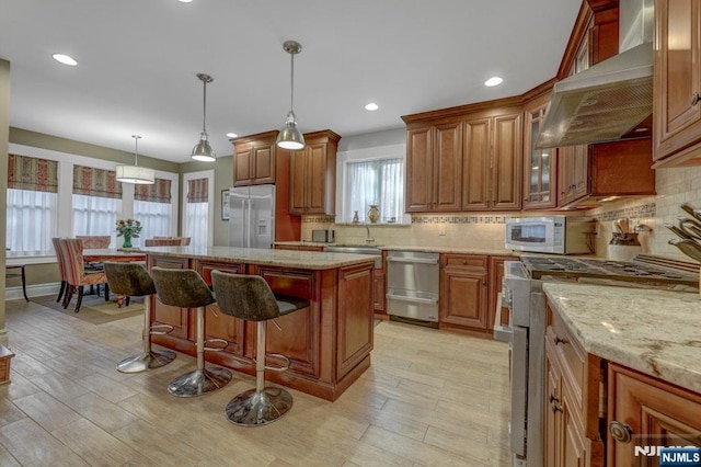 kitchen with a kitchen island, wall chimney range hood, a breakfast bar, brown cabinets, and appliances with stainless steel finishes