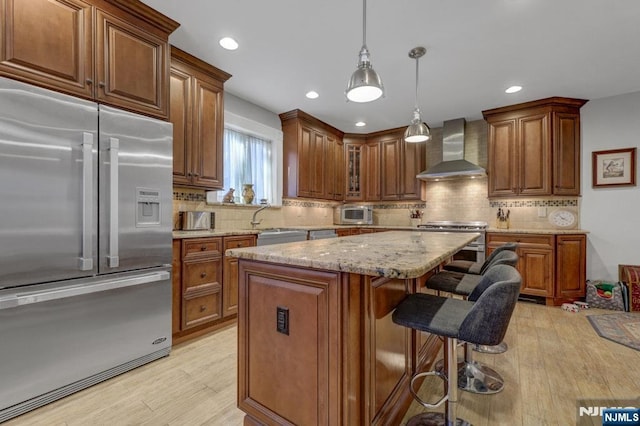 kitchen featuring light wood finished floors, a kitchen island, light stone counters, appliances with stainless steel finishes, and wall chimney exhaust hood