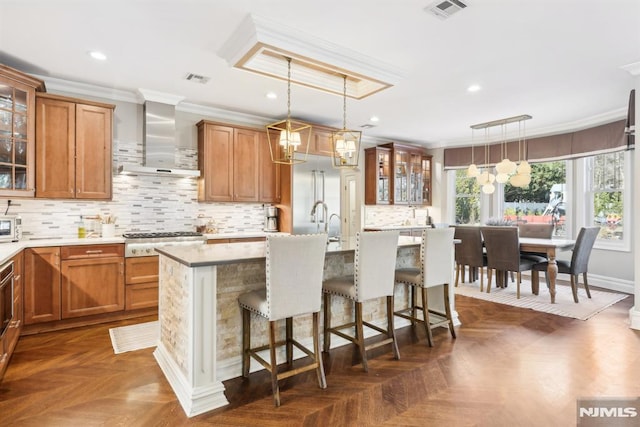 kitchen with a kitchen island with sink, hanging light fixtures, wall chimney range hood, and glass insert cabinets