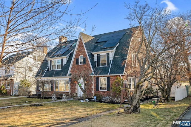 view of front of property with a shingled roof, brick siding, fence, roof mounted solar panels, and a front lawn