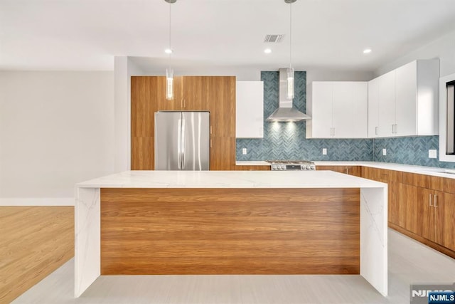 kitchen featuring a kitchen island, pendant lighting, stainless steel refrigerator, white cabinets, and wall chimney range hood