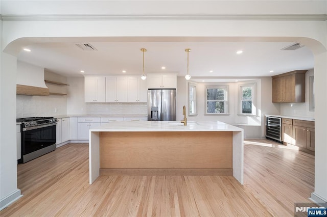 kitchen featuring an island with sink, wine cooler, white cabinets, hanging light fixtures, and stainless steel appliances