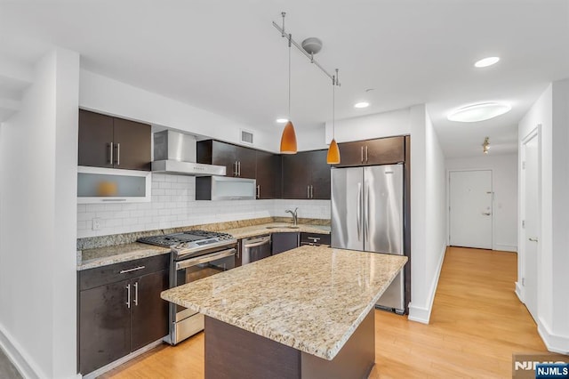 kitchen with appliances with stainless steel finishes, decorative light fixtures, a center island, dark brown cabinets, and wall chimney range hood