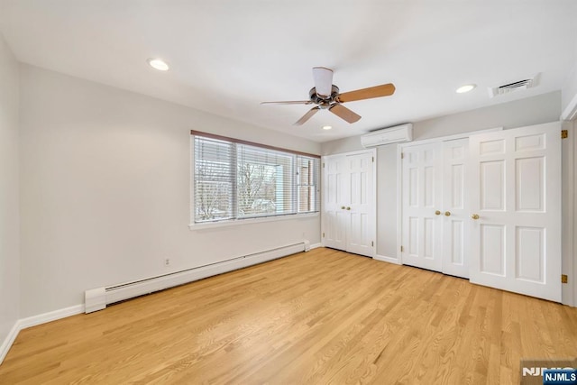 unfurnished bedroom with ceiling fan, a wall mounted AC, light hardwood / wood-style floors, baseboard heating, and two closets