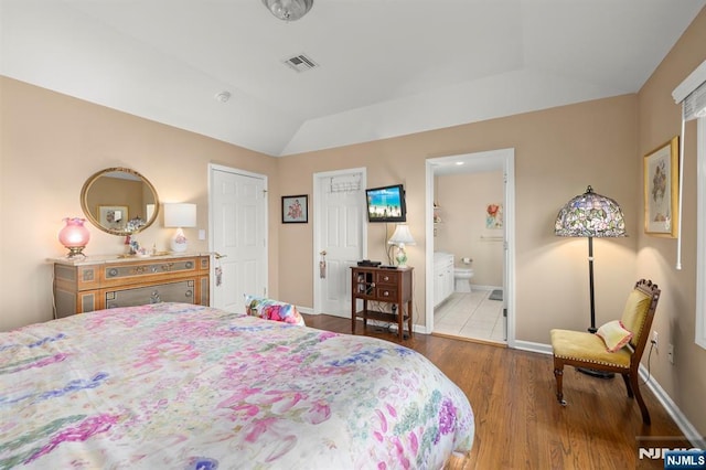 bedroom with light wood finished floors, baseboards, visible vents, lofted ceiling, and ensuite bathroom