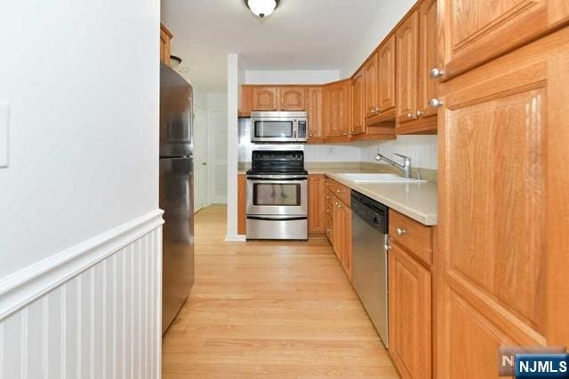 kitchen with light countertops, appliances with stainless steel finishes, brown cabinetry, wainscoting, and a sink