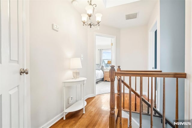 hallway featuring baseboards, visible vents, wood finished floors, an upstairs landing, and a notable chandelier