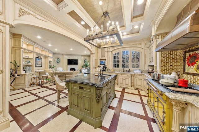 kitchen with custom exhaust hood, pendant lighting, crown molding, stainless steel gas cooktop, and a kitchen island with sink