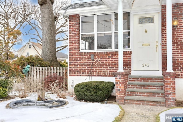 snow covered property entrance with brick siding, roof with shingles, and fence