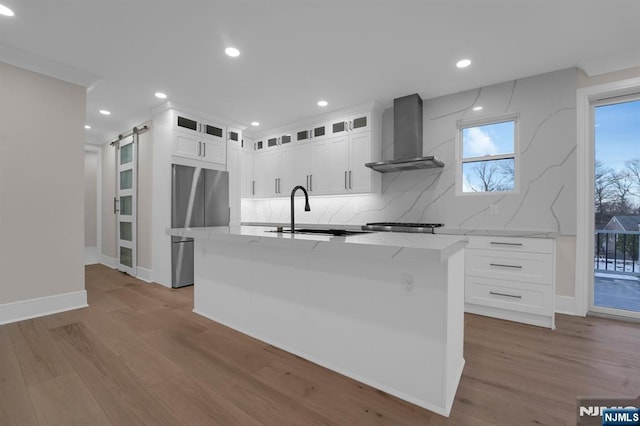 kitchen with a center island with sink, a barn door, white cabinets, and wall chimney range hood