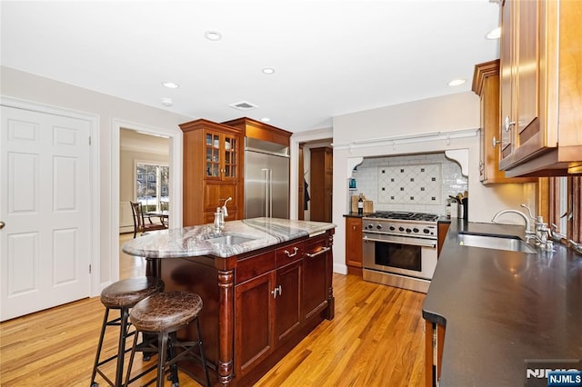 kitchen with light stone counters, premium appliances, a sink, a center island with sink, and glass insert cabinets