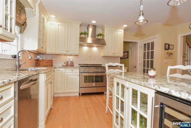 kitchen with a sink, light stone counters, wine cooler, appliances with stainless steel finishes, and wall chimney exhaust hood