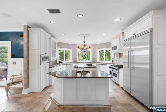 kitchen featuring a center island, custom range hood, hanging light fixtures, glass insert cabinets, and built in appliances