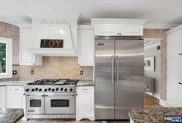 kitchen featuring premium appliances, white cabinets, crown molding, and dark stone countertops