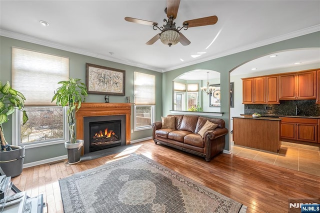 living area with baseboards, a glass covered fireplace, light wood-style flooring, ornamental molding, and ceiling fan with notable chandelier