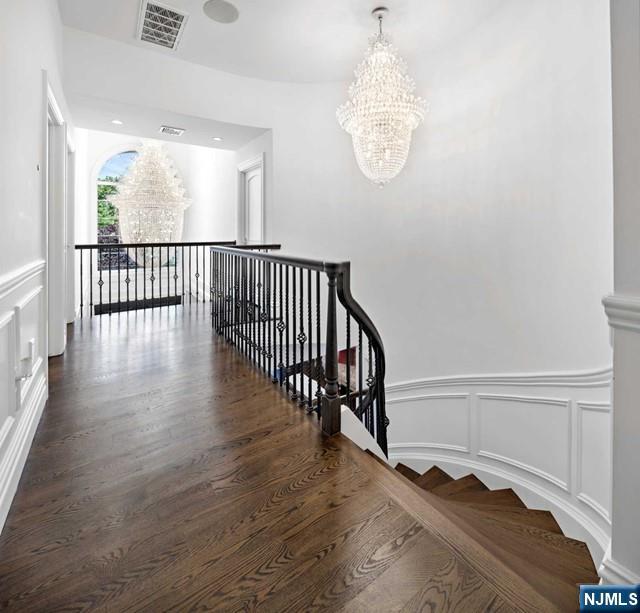 corridor featuring wood finished floors, visible vents, a decorative wall, a notable chandelier, and an upstairs landing