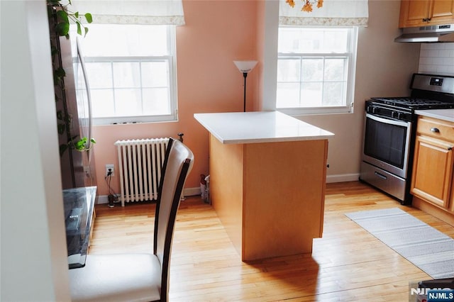 kitchen featuring light wood finished floors, radiator, light countertops, under cabinet range hood, and stainless steel range with gas stovetop