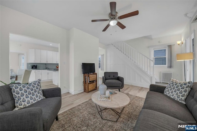 living room with radiator, light wood finished floors, ceiling fan, and stairs