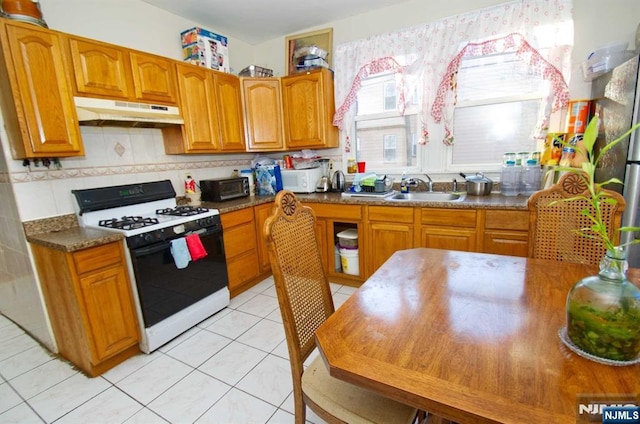 kitchen with brown cabinets, light tile patterned floors, gas stove, a sink, and under cabinet range hood