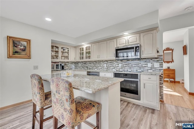 kitchen featuring light wood-style flooring, stainless steel appliances, a kitchen breakfast bar, light stone countertops, and glass insert cabinets