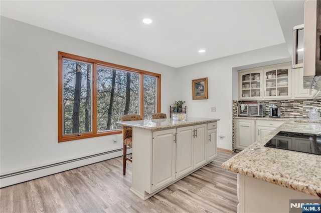 kitchen with light stone counters, baseboard heating, light wood finished floors, a kitchen bar, and glass insert cabinets