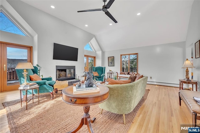 living area with high vaulted ceiling, a ceiling fan, light wood-style floors, baseboard heating, and a glass covered fireplace