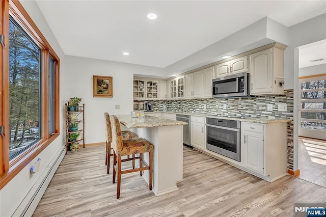 kitchen with a healthy amount of sunlight, appliances with stainless steel finishes, baseboard heating, light stone countertops, and glass insert cabinets