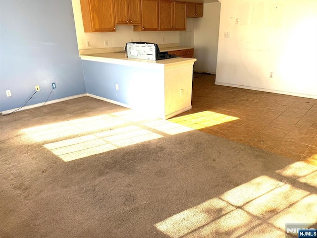 kitchen featuring light carpet, a peninsula, baseboards, light countertops, and brown cabinetry